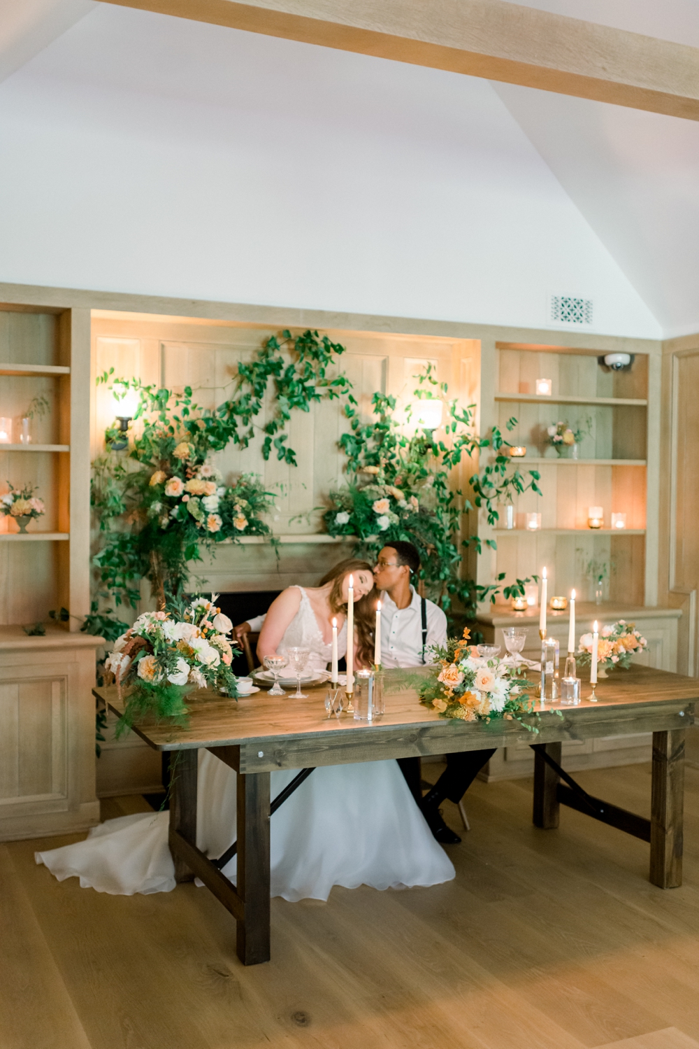 A bride leans on her groom's shoulder as he kisses her head at their head table with greenery