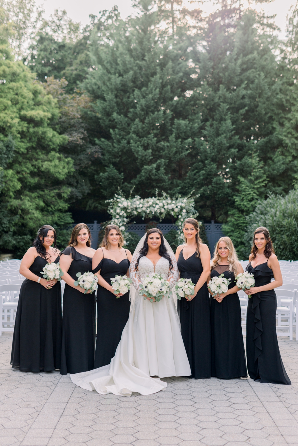 A bride stands in her outdoor ceremony location with her bridesmaids in black dresses