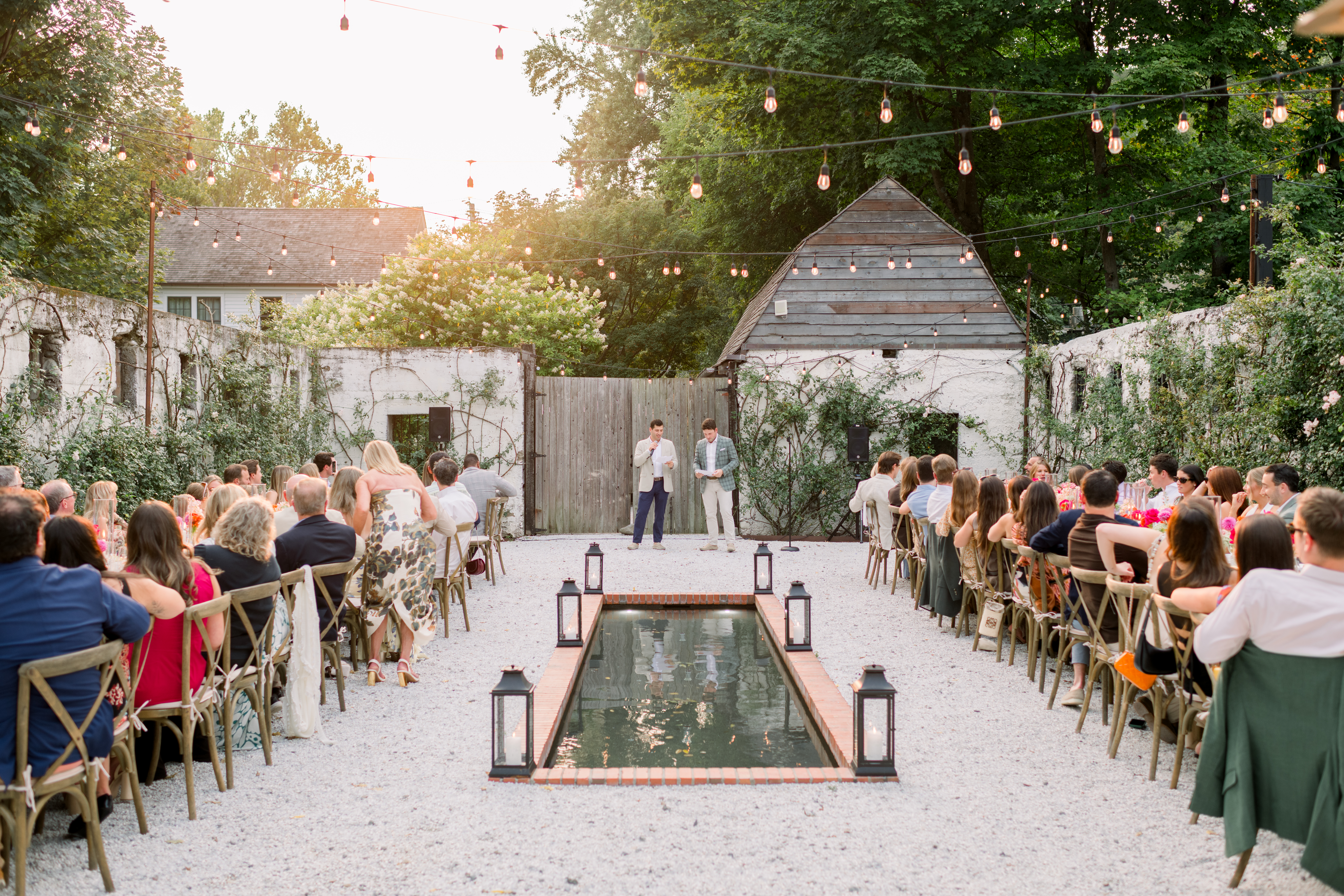 Wedding guests sit at decorated reception tables in a garden listening to speeches from wedding rehearsal dinner ideas
