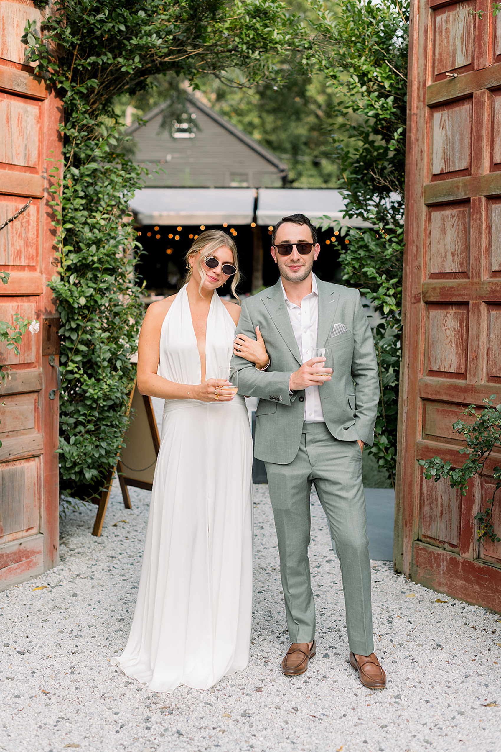 A bride in white and groom in green stand in the entrance to a garden wedding rehearsal dinner after finding ideas with sunglasses