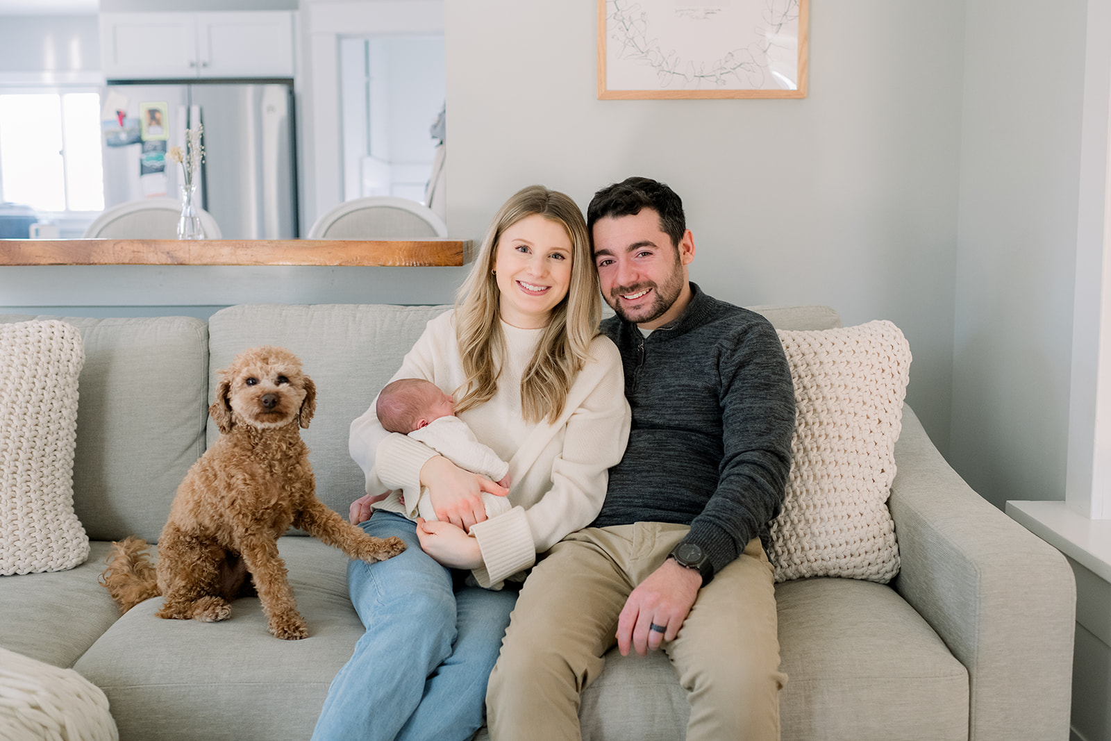 New mom and dad sit on a couch smiling and holding their newborn as their small brown dog puts a paw on mom's leg after listening to newborn photo session tips