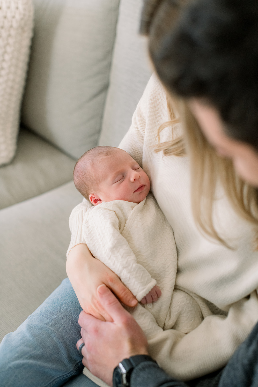 A look down at a newborn baby in a white onesie sleeping in mom's arms on a couch