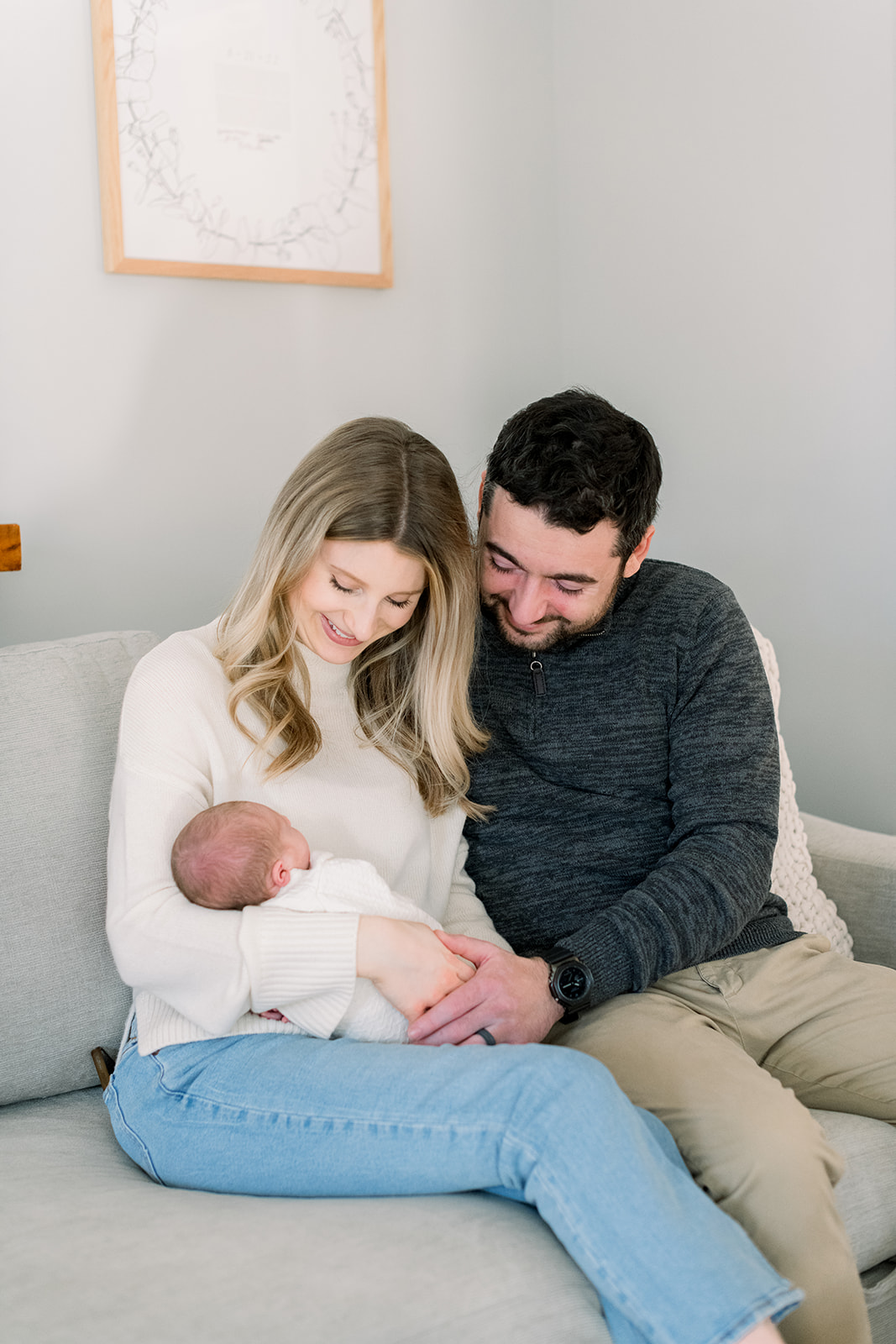 Smiling new mom and dad sit close on a couch looking down at their newborn in mom's arms