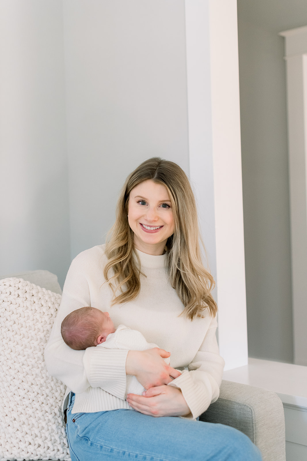 A happy new mother sits on a couch smiling in a cream sweaters holding her sleeping newborn