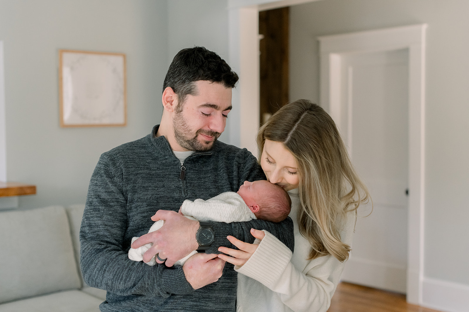 Mom kisses her sleeping newborn in dad's arms while standing in the living room