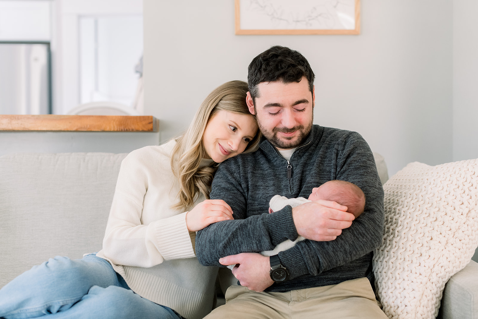 After learning some newborn photo session tips, a mom and dad sit on a couch snuggling while cradling their sleeping newborn