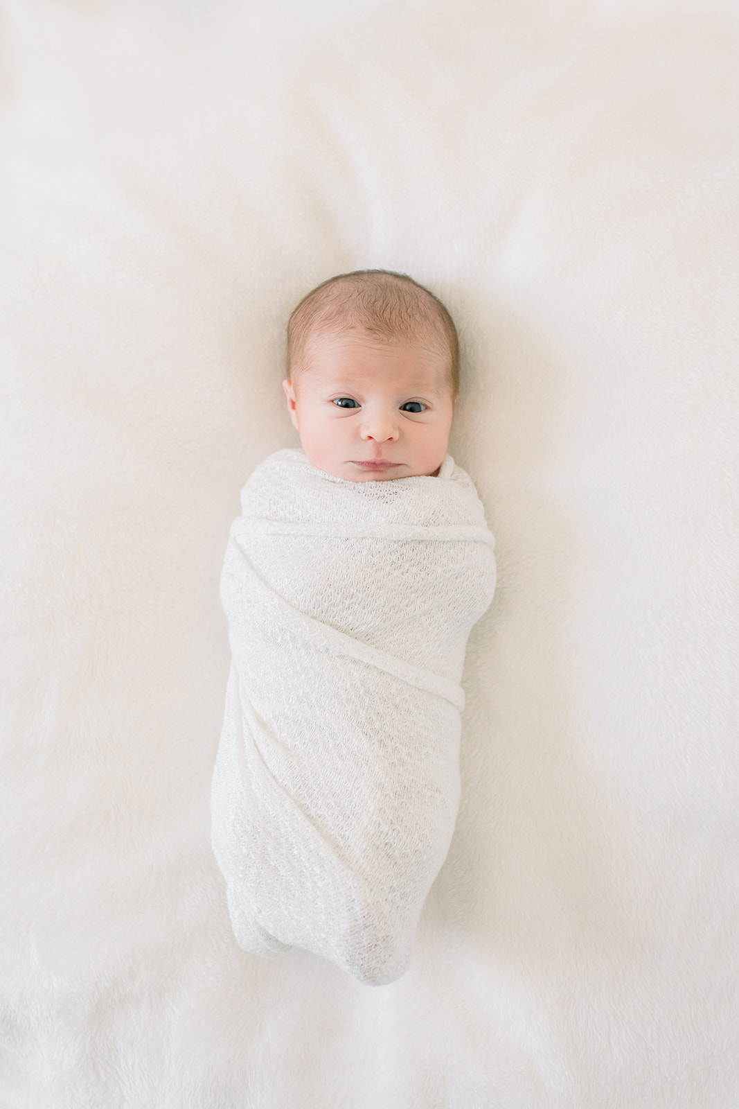 A look down at a newborn laying swaddled in a white crib with eyes open
