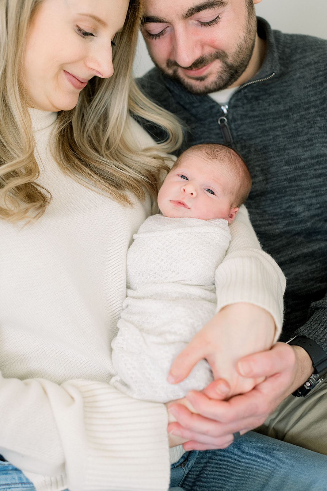 A newborn baby opens his eyes while swaddled in mom's arms beneath dad after listening to newborn photo session tips