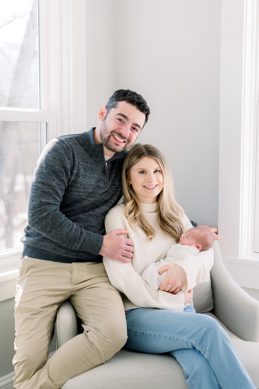 New parents in cream and grey smile while sitting in a nursing chair holding their newborn in a corner between windows