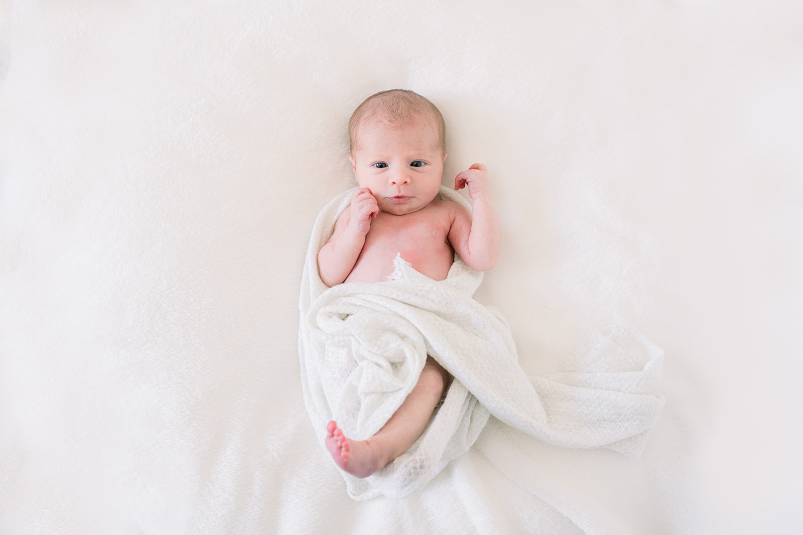 A baby loosely wrapped in a white blanket lays on his back in a crib awake thanks to newborn photo session tips
